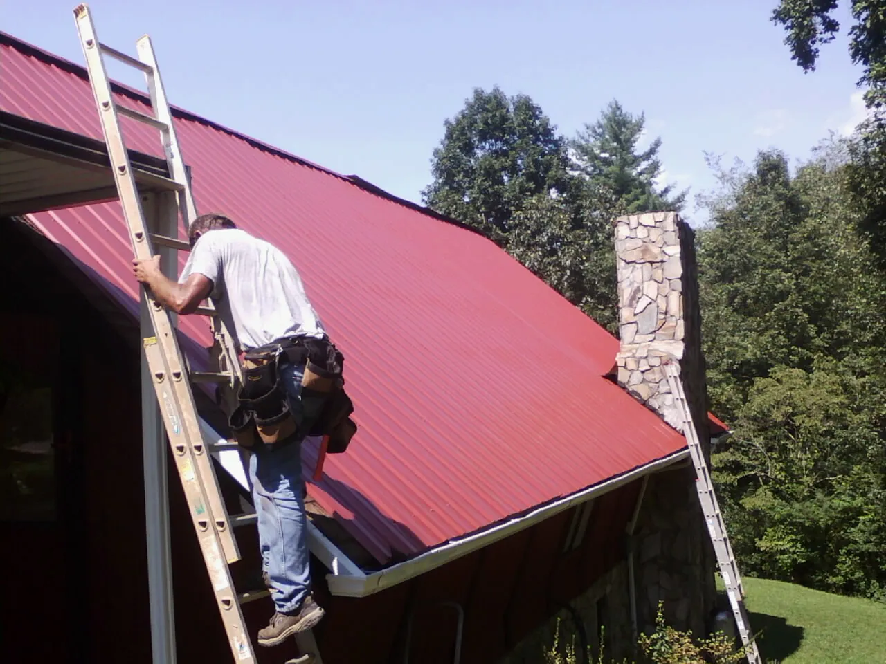 Roofer on a ladder beside a red metal roof and stone chimney — Soffit & Fascia Repair by Roofing Brothers in Micanopy